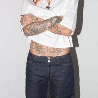 Person wearing a white shirt and dark jeans with tattoos on a plain background