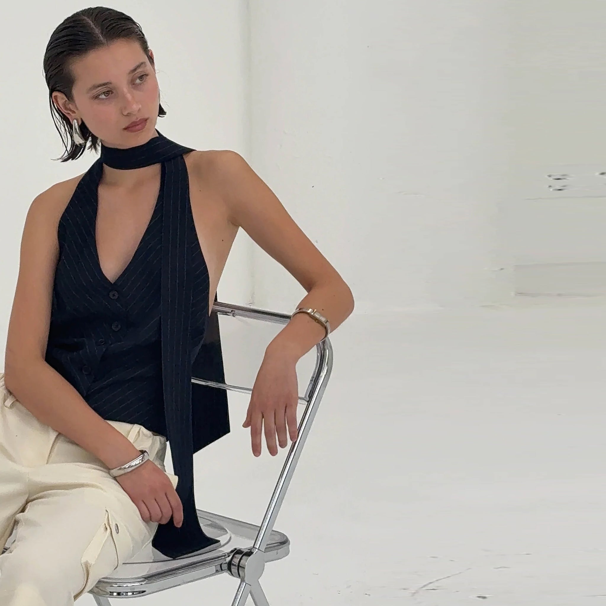 Woman sitting on a chair wearing a navy halter top and white pants against a white background