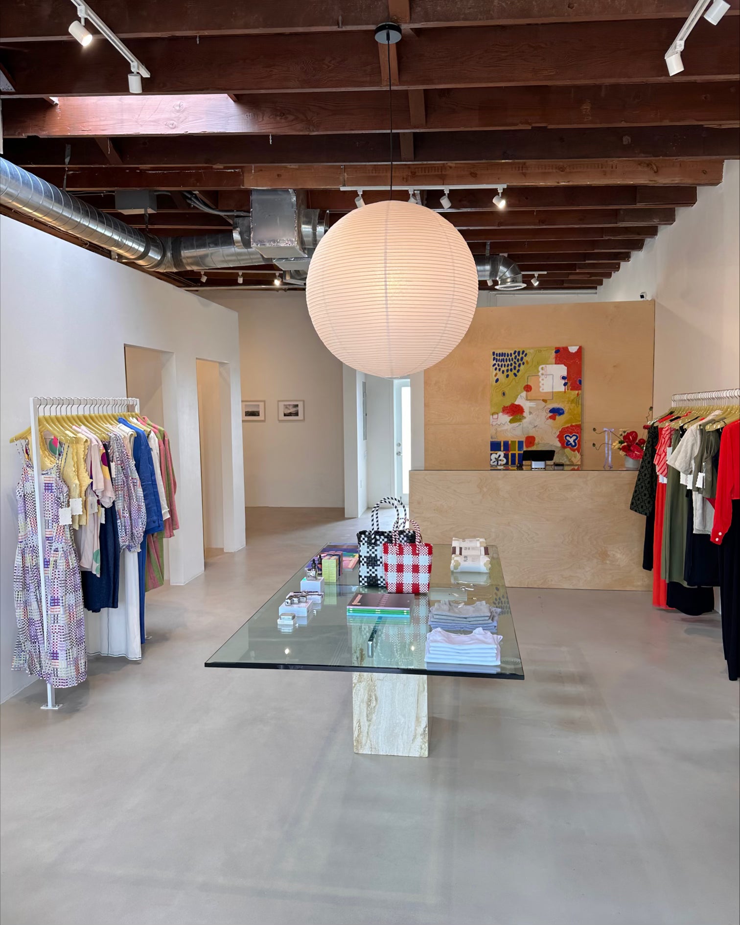 storefront with large glass travertine table and a large paper lantern pendant light surrounded by brightly colored clothes on racks